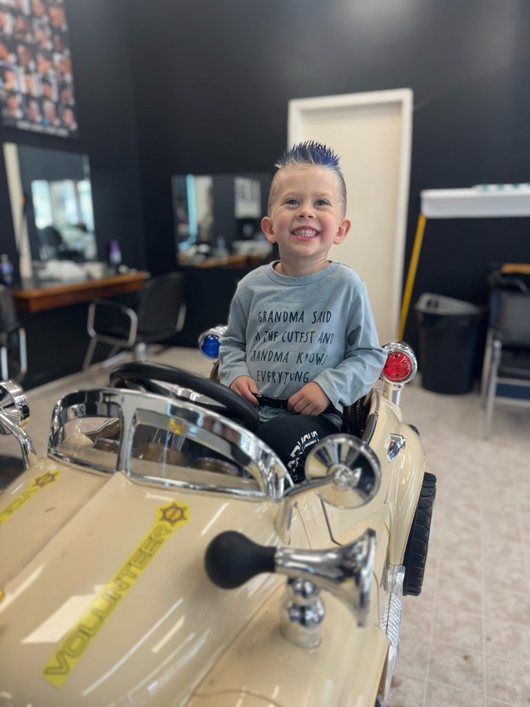 A smiling child with a fresh haircut sitting in the car-shaped barber chair